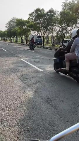 The video shows a view from a motorcycle as it travels down a road. A person on another motorcycle is in front and to the left of the camera. Trees line the right side of the road, and a truck is visible in the distance. The road surface appears cracked and uneven.