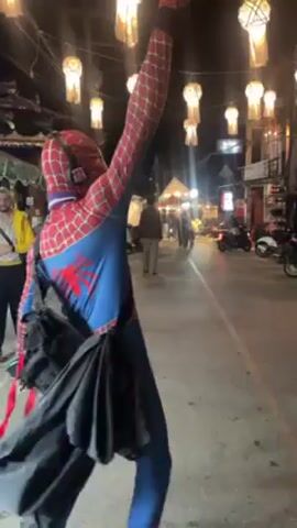 The video shows a person in a Spider-Man costume on a street at night. They have their arms raised in the air, wearing headphones, a black bag, and holding a black cape. A person in a black shirt and grey shorts approaches and gives the Spider-Man person a high five.  A string of lantern-like lights hangs above the street. There are people walking around and parked motorcycles.