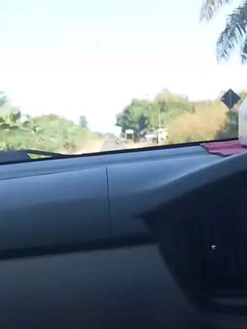The video shows a view from inside a car driving on a rural road. The dashboard of the car is visible in the foreground, with a red cloth and a clear plastic bottle on it. The road ahead is paved and appears to be a two-lane road. On either side of the road, there are trees, vegetation, and some small buildings or structures. There are also red and white striped posts along the side of the road. In the distance, a motorcycle is visible on the road. The sky is bright and clear, suggesting it is daytime. The camera appears to be mounted inside the car, looking forward through the windshield.