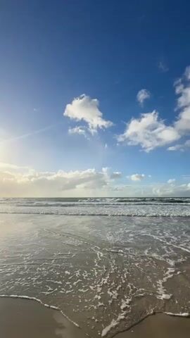 A vertical video captures a serene beach scene under a vibrant blue sky dotted with fluffy white clouds. The sun casts a gentle glow on the left side of the frame. Gentle waves roll onto the wet sand, leaving foamy trails as they recede. The ocean stretches to the horizon, meeting the sky. There are no people or objects visible in the frame.