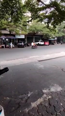 A short video shows a street scene with buildings and trees. Several cars and motorcycles are parked along the side of the road. A person walks near the parked cars, and a motorcycle is seen driving down the street. The ground in the foreground is paved with hexagonal tiles, and a drainage grate is visible. The overall impression is of a moderately busy street, possibly in a town or city.
