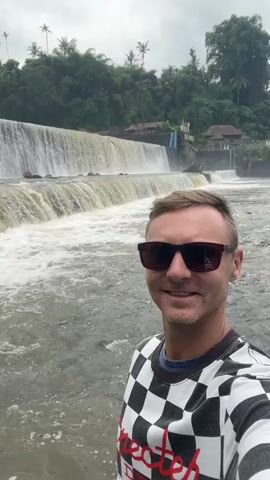 This is a vertical, first-person video selfie featuring a man standing in front of a wide waterfall. The man is wearing sunglasses and a black and white checkered long-sleeved shirt with some red text visible on the front. He is smiling at the camera. The waterfall is a broad, tiered cascade with murky, churning water flowing over it. The setting appears to be a tropical environment, with lush green trees and palm trees visible on the hillside behind the waterfall. There are some buildings and structures associated with the waterfall, including what looks like a small concrete dam or control structure and a building on the right side of the waterfall. The sky is overcast, suggesting a cloudy day, possibly with recent rain. The overall mood is relaxed and happy, with the man appearing to enjoy the scenic location. The video is set in Klungkung, Indonesia, implying a tropical, potentially humid climate.