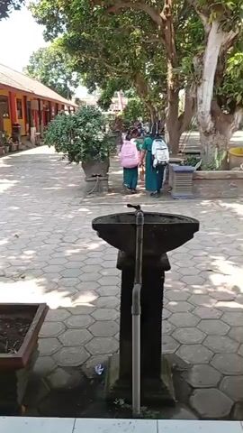 The video shows a stone washbasin with a faucet in the foreground. In the background, two young girls wearing green dresses and carrying backpacks are walking away from the camera down a paved path lined with trees and bushes. The girl on the left has a pink backpack, and the girl on the right has a white backpack with drawings on it. They are walking towards a building with a red roof. The scene appears to be outdoors in a schoolyard or similar public area.