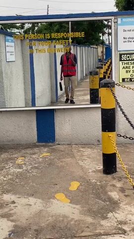 This video captures a person in a red safety vest and khaki pants walking towards a reflective surface. The reflective surface appears to be a large mirror or polished metal wall, likely part of a building's exterior. The person is holding a dark object in their left hand and is looking at their phone, which is held up in front of their face, suggesting they are taking a selfie or recording a video of their reflection.

The reflection reveals a yellow text that reads: "THIS PERSON IS RESPONSIBLE FOR YOUR SAFETY IN THIS BREWERY". This indicates the setting is likely within or adjacent to a brewery facility. The background visible in the reflection shows a walkway lined with alternating black and yellow striped bollards, some with chains connecting them, and greenery including trees and potted plants. The overall scene suggests an industrial or operational area within the brewery.

The ground in the foreground of the video is concrete with several bright yellow splotches, possibly spilled liquid or paint. The lighting suggests it is daytime, with overcast or diffused sunlight, as there are no harsh shadows. The mood appears casual, with the individual focusing on their phone. The location context of Kinkinau, Nigeria, provides a geographical anchor to the visual information.