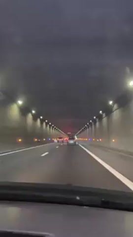 The video shows a car driving through a lit tunnel. There are evenly spaced lights on the tunnel walls, and other cars visible in the distance.  The driver's perspective is from inside the car, looking through the windshield.  The road is clearly marked with white lines.  A silver SUV passes in the adjacent lane. The tunnel walls appear to be a grey color.