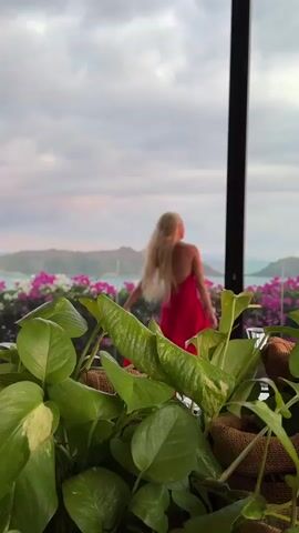 A woman with blonde hair is standing on a balcony overlooking a body of water and hills. She is wearing a red dress. In the foreground, there are large green leaves of a plant. The sky is cloudy.