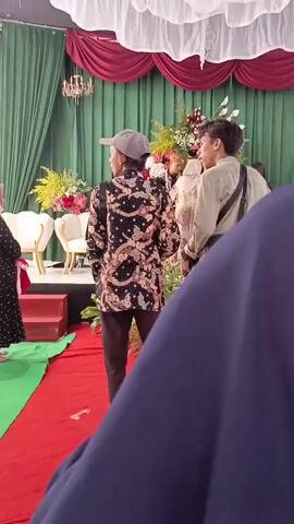 The video begins with a view of a stage decorated with green and burgundy drapes and white fabric at the top. There are floral arrangements and white chairs on the stage. A person in a black patterned shirt and a cap is standing in the foreground, facing away from the camera. To the right, a young man in a light-colored shirt with a bag slung across his chest is also visible. Further back on the stage, a woman in a headscarf is standing, and there are more people in the background. The scene appears to be set up for an event, possibly a wedding or a formal ceremony. As the video progresses, the camera shifts focus and shows more of the audience and the stage. Several people in traditional attire are on the stage, and one person is holding a microphone, seemingly addressing the attendees. A young child is in the immediate foreground, looking towards the stage. The lighting suggests an indoor setting, possibly a hall or a tent. The overall atmosphere seems to be one of solemnity and occasion.