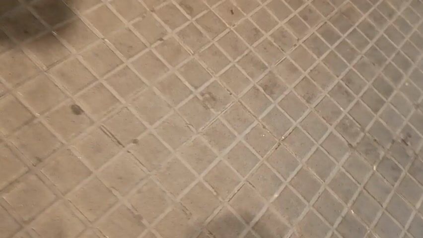 A person wearing white sneakers, black pants, and a dark top walks along a paved area. Their shadow is visible on the pavement. The person walks past a traffic light with a green walking man symbol. The video is shaky and appears to be shot from a low angle.
