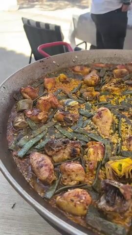 Here's a description of the video:

The video shows a large paella dish filled with rice, chicken pieces, green beans, and artichoke hearts. The food is cooked and ready to be served. The paella pan is placed on a table outdoors. There are people standing around the table, seemingly waiting to enjoy the meal. The setting appears to be a pleasant outdoor area, possibly a patio or garden, with trees and other tables visible in the background. The video offers a close-up view of the dish, then widens to reveal the surrounding social gathering.