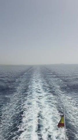 A wide shot captures a wake trailing behind a boat at sea. The water is a deep blue and is churned up into white foam behind the vessel, forming a distinct path through the ocean. In the foreground, a flag with red, yellow, and possibly brown stripes can be seen attached to a pole on the right side of the boat. The horizon is visible in the distance, with a hazy sky above. The overall impression is one of movement and travel across a vast body of water.