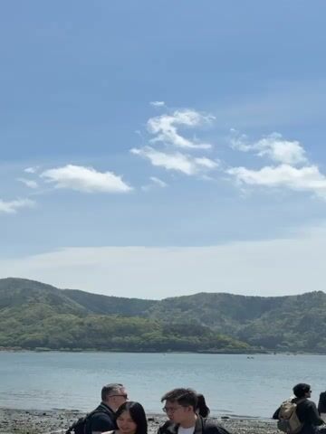 The video shows a scenic outdoor landscape featuring a lake, mountains, and a clear blue sky. In the foreground, a group of people are standing by the water's edge, seemingly enjoying the view. The camera pans from left to right, revealing a wide expanse of water, likely a lake, with forested mountains rising across the opposite shore.

As the camera continues to pan, the iconic snow-capped peak of Mount Fuji comes into view on the right side of the frame. The sky is bright blue with scattered white clouds, indicating fair weather. The people in the foreground appear to be tourists or visitors, some looking out at the landscape. The overall atmosphere is peaceful and picturesque, capturing a beautiful day in Fujikawaguchiko, Japan. There is no visible text in the video.