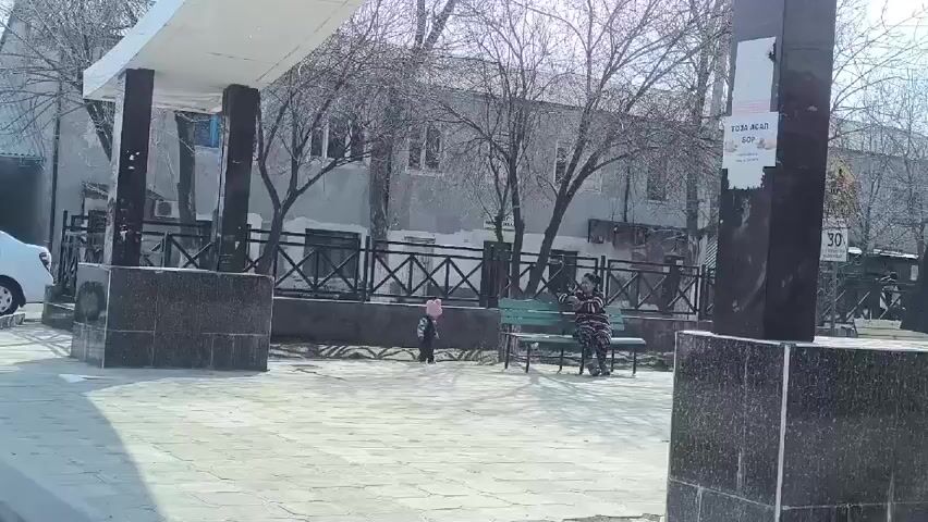 The video shows a small square in what appears to be a residential area. A woman sits on a green bench, possibly using a phone. A toddler in a pink hat stands nearby. As the camera shifts, three young men walk past under a small overhang. Bare trees are visible in the background, along with buildings of a muted color. The ground is covered in light-colored paving stones.  A sign is attached to a pillar next to the camera.