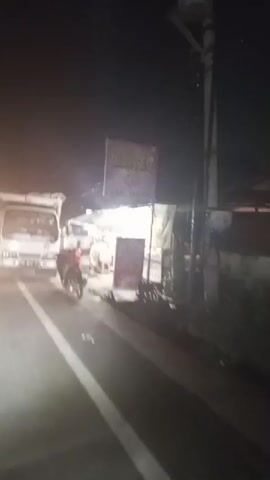 The video opens to a nighttime scene of a white truck with a tarp-covered bed parked on the right side of a paved road. A motorcycle is parked in front of the truck, and a person in a red shirt is standing near it. Another person is seen walking away from the motorcycle towards the truck. The truck has headlights on, illuminating the road. In the background, there are what appear to be food stalls with awnings and lights. A sign with some text is visible above the stalls. The road has white lane markings. The camera then pans to the side of the truck, showing its wooden bed. The scene continues with a view of the truck's side and then its rear as it appears to be moving forward, with lights from other vehicles visible in the distance on the road.