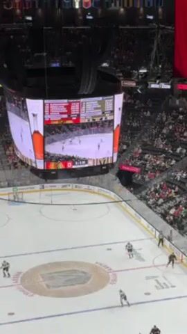 A high-angle shot shows a hockey game in progress in a large indoor arena. Players are on the ice, illuminated by bright lights. Overhead, a large, multi-sided video board displays the game footage, statistics, and advertisements. The arena is filled with spectators. The ice rink features markings and logos. A referee is visible on the ice. The score is 0-4.