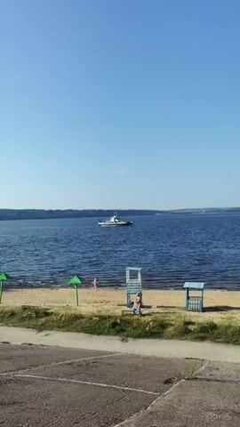 The video shows a calm, wide body of water under a clear blue sky. A white boat with a dark superstructure is visible in the distance, sailing on the water. The foreground features a sandy beach with a few beach chairs, a small blue and white shelter, and green umbrellas. There are a couple of people on the beach, appearing to be relaxing. The water's edge is visible, with small waves lapping at the shore. The ground in the very front of the frame appears to be paved or made of concrete.