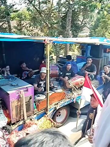 The video shows an outdoor scene with a truck modified to carry musical equipment and performers. A blue tarp covers the top of the truck bed, which is filled with large speakers, drums, and other sound equipment. Several individuals are seated or standing on the truck, some appear to be musicians, while others are observing or participating in the activity. One person is holding an Indonesian flag. In the foreground, a person in traditional attire, possibly a performer, is visible from behind. The background consists of trees and some buildings, suggesting a rural or semi-urban setting. The overall atmosphere appears to be festive, possibly related to a performance or celebration.