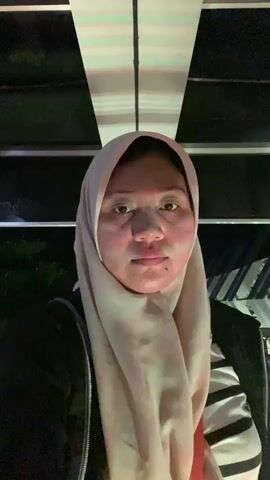 The video displays a woman wearing a light-colored hijab, a black jacket, and a black and white striped shirt. She is also carrying a bag with an orange strap. The scene appears to be outdoors at night, with green lights illuminating the background and a striped awning. There's a metal fence in the foreground, and what looks like a building or structure with white beams overhead. The woman is initially facing the camera and smiling, but then turns to her left, and her face becomes obscured by a mask. The activity seems to be a casual setting, possibly a public space or an event, given the lighting and background elements. There is some text visible on the fence in the background that reads "DON'T STOP TALKING ABOUT..."