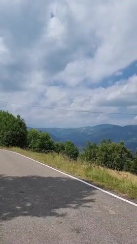 The video pans across a scenic view of rolling hills and mountains under a partly cloudy sky. The foreground shows a paved road with a white line marking the edge. On the right side of the road, there is a grassy slope dotted with trees and vegetation. In the distance, layers of mountains fade into a hazy blue, suggesting a vast landscape. The sky is a mix of bright blue and fluffy white clouds. The overall impression is a peaceful, natural environment.