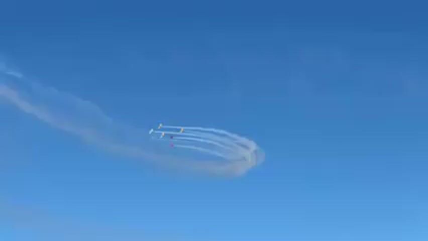 The video shows an aerial display featuring a group of planes performing maneuvers against a clear blue sky. The planes are emitting white smoke trails, which create patterns in the air. The formation of planes is tight and coordinated, creating a visually appealing display. The planes loop around, leaving a circular smoke trail in the sky. A structural element briefly comes into view in the upper left corner of the frame, obstructing the view of the sky.