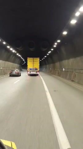 A first-person perspective video shows a vehicle driving through a well-lit tunnel. Ahead, a yellow semi-truck is visible, with a black car to its left. The tunnel walls are made of concrete and are illuminated by rows of lights. The road surface is asphalt, with a white line marking the lanes. The vehicle appears to be moving at a steady pace.