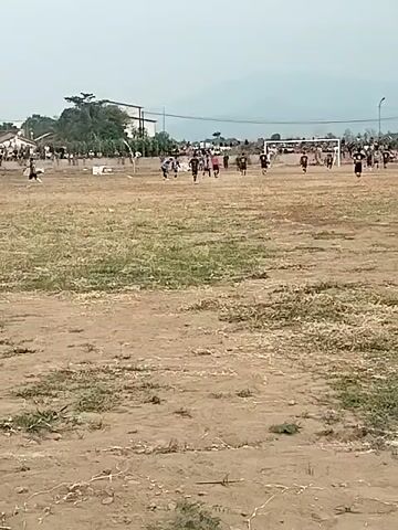 A wide shot captures a group of people on a dirt soccer field. The field is mostly bare with patches of dry grass. In the background, there is a soccer goal with a net, and several players are visible on the field. To the left of the field, there are some buildings and trees. In the distance, hills or mountains can be seen under a hazy sky. The overall scene suggests a casual soccer game or practice taking place in an open, undeveloped area.