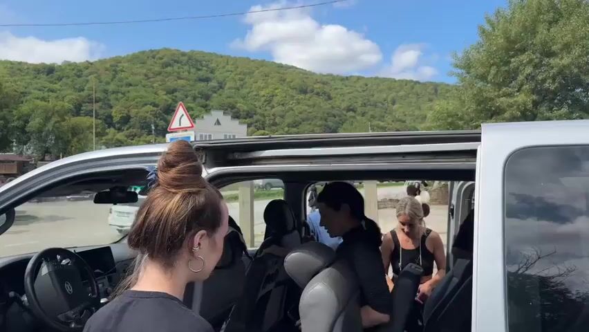 The video shows a group of people gathered around the open side door of a silver van. The van's interior, including the driver's seat and passenger seats, is visible. Several people are inside and around the van, appearing to be loading or unloading items. A woman in the foreground is seen from behind, with her hair tied up in a bun. In the background, there is a green, tree-covered hillside, a road with a traffic sign, and a building. The sky is blue with some clouds. The scene appears to be outdoors in daylight.