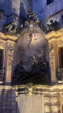The video displays an evening view of a crowded outdoor or semi-outdoor dining/social area. The background features a tall, elaborate stone architectural structure, possibly part of a building facade or a large fountain. This structure is composed of multiple levels: the uppermost part shows decorative carvings and figures, below which is a recessed arch containing a detailed stone relief depicting several figures, including what appears to be a man and a woman in a narrative setting, with winged cherubs above. Beneath this relief is a large, rectangular, un-inscribed stone plaque. The entire stone structure is illuminated by warm, upward-facing artificial lights. In the lower and mid-ground, numerous individuals, predominantly young adults, are seated at white tables arranged in rows. They are observed conversing, eating, drinking from glasses, and interacting with mobile phones. On the right side of the frame, a person is playing a brass trumpet. The camera executes a slow, slight zoom-out motion, widening the view of the gathering.
