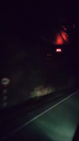 The video shows a nighttime view from inside a vehicle traveling on a road. The road is dark with a white line marking the lane. To the left of the road is a steep, rocky embankment covered in vegetation. In the distance, there are a few blurry lights that appear to be from buildings or other vehicles. There is a digital display in the upper right corner of the frame showing the number 35.5. The overall impression is one of traveling through a dark, rural area at night.