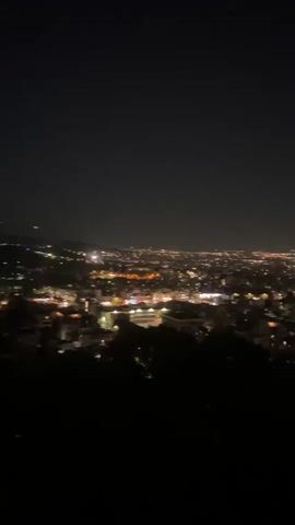 This is a vertical, slightly shaky, handheld video taken at night overlooking a city. The sky is black and clear, with a few distant lights appearing as small white dots. Below the sky, the city stretches out with countless lights twinkling across the landscape. The lights are primarily yellow and white, illuminating buildings and streets. In the foreground, the silhouette of dark trees and foliage obscures the lower part of the frame, creating a stark contrast with the illuminated city beyond. Towards the right side of the foreground, a metallic chimney pipe is visible. The overall impression is of a sprawling urban area at night, viewed from a high vantage point.