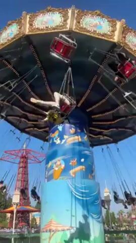 The video shows a spinning swing ride at what appears to be a Disney theme park. The ride, which features seats suspended by chains from a rotating canopy, is in motion with passengers riding in the swings. The center of the ride is decorated with Disney cartoon characters and painted in a blue gradient that mimics the sky. In the background, a red tower is visible against a clear blue sky. The camera angle is low, looking up at the ride.