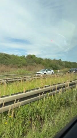 A video shows a view from a vehicle window. In the foreground, tall green grass and plants grow abundantly. In the middle ground, a railway track runs horizontally. Beyond the tracks, a road is visible with several cars driving on it. The landscape is hilly and covered with greenery, with a cloudy sky above.