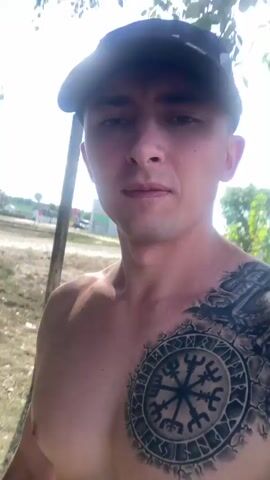 A man wearing a black baseball cap and no shirt stands outdoors. He has a large tattoo on his chest and left shoulder. The man flexes his bicep. The background shows grass, a pole, trees, and a building.