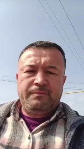 A man with short, dark hair, a beard, and wearing a purple shirt and a plaid jacket looks up at the camera.  He brings his hand into the frame and makes a peace sign.  The background is a bright, clear sky with power lines.