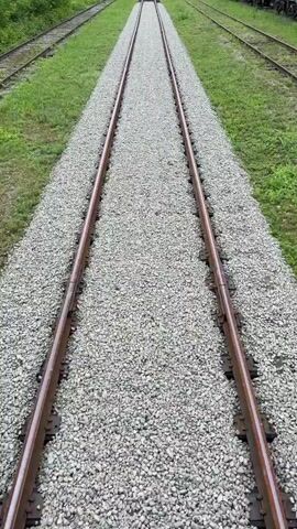 The video shows a high-angle shot of two parallel train tracks. The tracks are made of metal rails and are laid on a bed of grey gravel. Green grass grows on either side of the gravel bed. The tracks recede into the distance, appearing to converge at a vanishing point. There are no people or vehicles visible on the tracks. The overall environment appears to be outdoors, possibly in a rural or undeveloped area. The lighting suggests it is daytime.