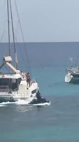 The video shows someone riding a black personal watercraft (PWC) or jet ski on the open water. The water is blue with patches of turquoise, and the sky is pale blue and hazy.  The PWC kicks up a spray of water as it moves. In the background, there are two large white sailboats.  People are visible on one of the sailboats. The PWC moves between the sailboats.