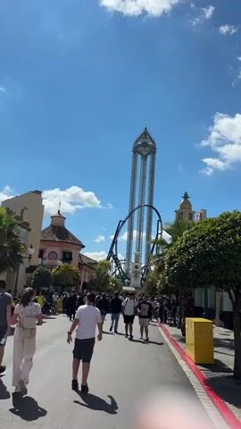 The video shows a wide street scene, likely a theme park, on a bright, sunny day with a blue sky and scattered white clouds. A crowd of people are walking in the same direction down the street. Several architectural styles can be observed in the buildings lining the street, including a Moorish-style structure with a domed roof, a building with a tall cross on its roof and decorative elements, and an ornate structure with red accents and Asian-inspired decorations.

A very tall roller coaster structure is visible in the background. The street has a gray paved surface with a red painted stripe at the edge. A yellow trash can is positioned along the street. Several green trees and palm trees are interspersed among the buildings.