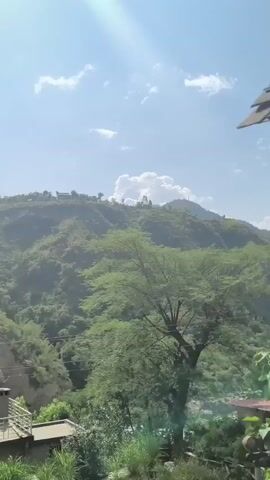 The video shows a panoramic view of a mountainous landscape under a bright, sunny sky with scattered clouds. The terrain is covered in lush green vegetation, with terraced farming visible on some slopes. In the foreground, there are some buildings and structures, including a house with a balcony and a water tank on its roof. The camera appears to be moving slowly, panning across the scene, capturing the natural beauty of the area and the human presence within it.
