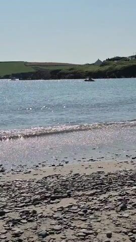 The video shows a rocky beach with calm, blue water. In the distance, there is a green hill with a few houses on it. The sun is shining, and the sky is clear. The waves are gently lapping at the shore, creating a sparkling effect on the water. There are no people visible on the beach or in the water.