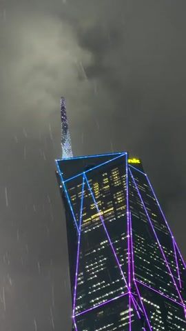 This media file shows a close-up, low-angle shot of a modern skyscraper at night during a rain shower. The building is tall and appears to be made of dark glass. It is illuminated by a striking display of neon lights that outline geometric patterns along its edges. These lights are primarily in shades of blue and purple, creating a vibrant contrast against the dark facade and the stormy sky.

The skyscraper has a unique, pointed spire at its very top, which is also decorated with sparkling lights, giving it a crystalline appearance. Many windows on the building are lit from within, suggesting that it is occupied. On the upper section of the building, yellow text is visible, spelling out "Maybank," indicating that this is likely the Maybank Tower in Kuala Lumpur, Malaysia.

The weather is clearly rainy, with visible streaks of rain falling from the dark, overcast sky. The overall mood is dramatic and perhaps a little eerie due to the low light, rain, and the imposing, illuminated structure. The focus is solely on the architectural marvel and the atmospheric conditions, with no people or other objects visible in the frame. The time of day is unequivocally night.