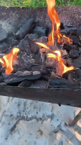 The video shows a close-up view of charcoal burning in a grill. Flames are visible, with some smoke rising from the coals. The grill appears to be made of metal and is placed outdoors, with grass and some yellow flowers visible in the background. The ground beneath the grill is a light-colored, possibly concrete or stone surface with some white debris. The camera angle is looking down into the grill. There are no people visible in the video. The scene suggests preparation for grilling or cooking.