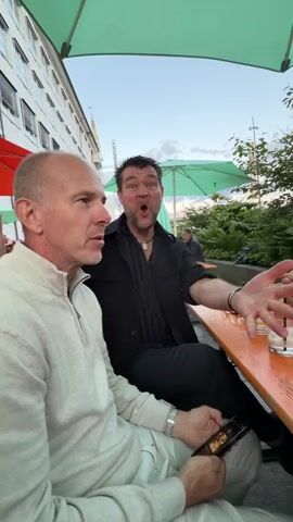 Three men and one woman are seated around an orange picnic table outdoors. The man in the foreground on the left is wearing a cream-colored sweater and is looking to his left. A second man, with dark hair and a black shirt, is in the center, looking at the camera and speaking animatedly with his hands raised. A third man, with gray hair and a green shirt, is seated opposite the first man and is looking towards the camera. A woman with glasses and a white sweater is on the far right of the frame, holding a glass and smiling. There are drinks on the table, along with a pack of cigarettes and a jar. The scene appears to be under a green umbrella, and there is greenery in the background. The time stamp at the beginning is 00:00 and it ends at 00:04.
