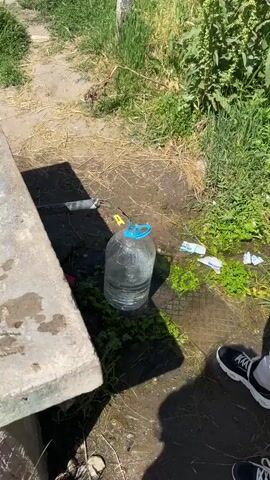 A clear plastic jug with a blue handle sits on the ground next to a concrete bench. A metal pipe extends from the left side of the frame, pouring water into the jug. The surrounding area is a mix of grass, dirt, and sparse vegetation. A pack of "MICKO" cigarettes lies discarded nearby. A person wearing black and white shoes stands to the right, with only their feet visible. The scene is illuminated by sunlight, casting shadows across the ground. The area appears to be slightly wet, with water accumulation around the jug.