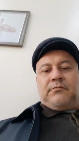 A middle-aged man with a salt-and-pepper beard and a black cap is looking at the camera. He is wearing a dark sweater with a brown collared shirt underneath, and a navy blue jacket. He looks up and to the side momentarily. The background is a plain white wall with a framed picture hanging on it. There is the sound of children crying in the background.