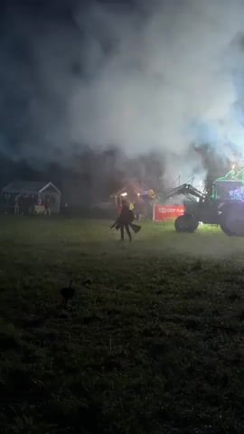 A low-angle night shot shows a grassy field with a person on the left foreground holding two fire-breathing torches. They move towards the camera, exhaling flames. Behind them are several tents and a tractor, which is also breathing fire. The tractor also has fireworks shooting up from its front. In the background, white smoke billows from the ground, obscuring part of the tractor.