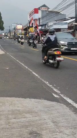 The video shows a road in an urban setting. Several people are riding motorcycles down the road. A yellow minivan also drives down the road. A store with a red and white facade and the name AMEN is visible in the background. There are power lines overhead. The sky is overcast.