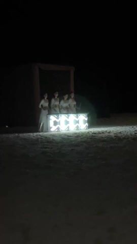 The video shows a group of four women in light-colored, perhaps white, dresses standing behind a small, illuminated structure in what appears to be a dark outdoor setting, likely a beach. The structure emits bright light with changing patterns. The women appear to be engaging with the structure, possibly playing instruments or interacting with the lights. Behind them is a structure with a rectangular opening, possibly a simple building or archway draped with fabric. The scene is dimly lit, with the light from the structure being the most prominent source of illumination. The camera is static, capturing the scene from a distance. The surface beneath the women and the structure looks like sand.