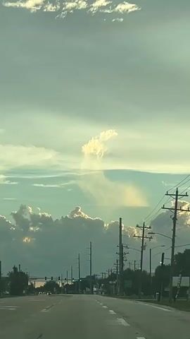 The video shows a view from a vehicle driving on a road. Overhead power lines are visible, along with telephone poles. The sky is cloudy with some sunlight breaking through. In the distance, there are cumulus clouds that appear to be illuminated from behind by the sun. A billboard reading "AMERICA'S LARGEST INJURY LAWYERS MORGAN & MORGAN" is also visible. There are trees and some buildings along the side of the road.