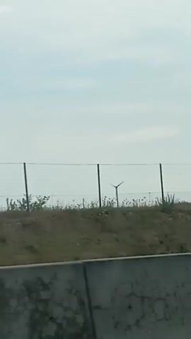 A video, likely captured from a vehicle, shows a landscape with a fence and wind turbines in the background. The foreground is dominated by a concrete barrier. The sky is overcast. The terrain behind the fence appears uneven, with some vegetation and rocks visible. Multiple wind turbines with blades are visible behind the fence, some appearing to be in motion. The camera moves, indicating a journey through the area.