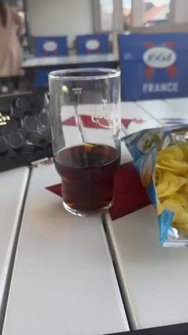 The video shows a table on a covered patio with a glass of dark liquid and a bag of potato chips. In the background, a person in a light-colored coat walks away from the camera. There are blue chairs with a "1664 FRANCE" logo visible. The sky above is blue with fluffy white clouds.