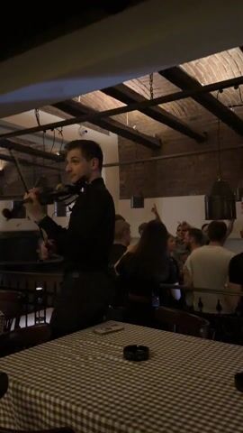 Inside a bar, a violinist in a black shirt and pants stands on a slightly raised platform, playing to a group of people. The crowd, a mix of men and women, is dancing and cheering, some with their arms raised. The bar has a rustic decor with exposed brick walls, wooden beams on the ceiling, and pendant lights. Tables with checkered tablecloths surround the dance area. An ashtray is placed on one of the tables in the foreground.